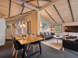 A spacious living area with a high ceiling, a dining table set for four, a sofa, and a television.