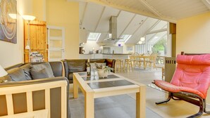 Amplia sala de estar con sofá, una mesa de comedor y una silla rosa. Se observa una cocina al fondo con tragaluz.