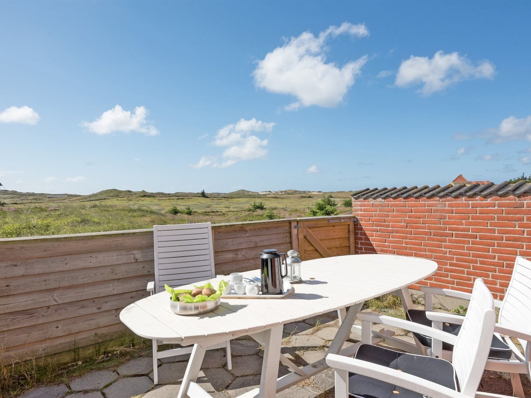 Une terrasse extérieure avec une table et des chaises blanches, un saladier rempli de fruits et un récipient en métal.