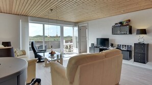 Ein modernes Wohnzimmer mit einer Couch, einem Couchtisch und einem Fernseher. Ein Balkon bietet Blick auf die Landschaft.