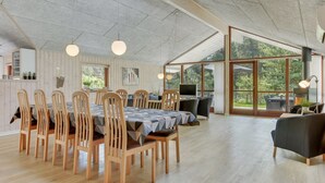 A spacious dining area with a long table and wooden chairs, a kitchen area with a TV, and large windows offering a view of the outdoors.