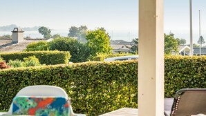 Vue depuis un espace extérieur abrité, avec une chaise et une table, donnant sur un jardin aménagé et un plan d’eau, avec des bâtiments visibles au loin.