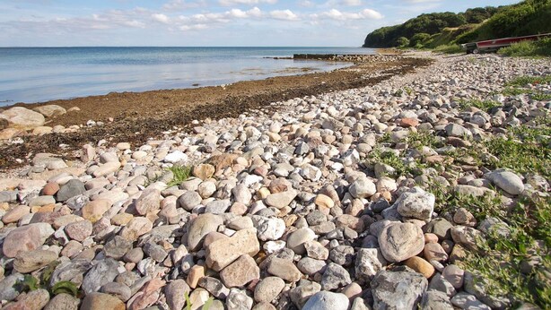 En stenstrand med utsikt över havet och ett grönskande kullområde i fjärran.