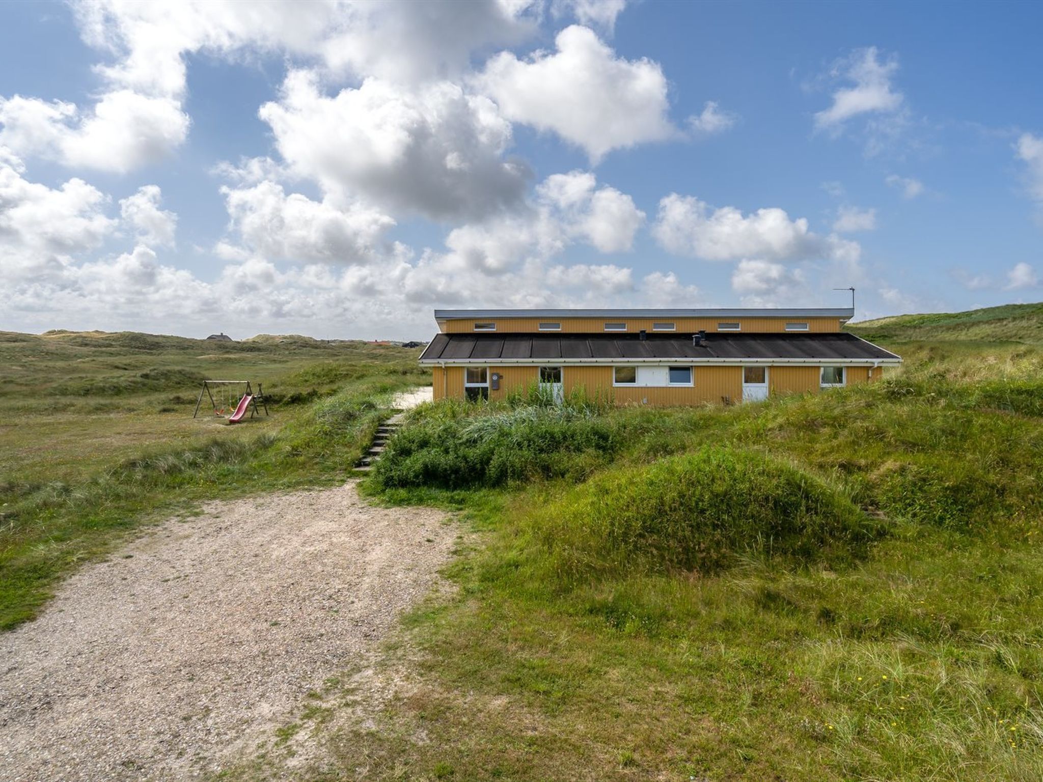 A two-story building with a gravel path leading to it, surrounded by grassy fields and dunes.
