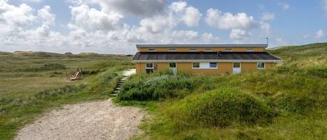 A two-story building with a gravel path leading to it, surrounded by grassy fields and dunes.