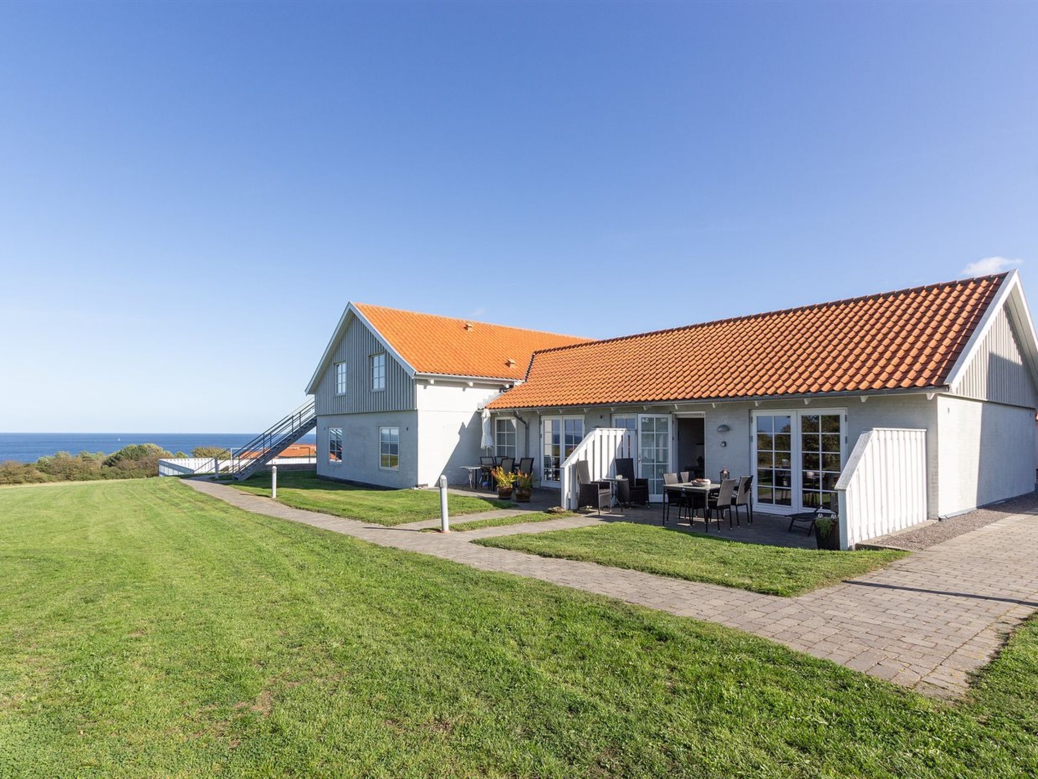 Una casa de dos pisos con techo de tejas rojas, un patio con muebles de exterior y vista al mar.
