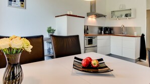 Eine moderne Küche mit weißen Schränken, einer Edelstahl-Dunstabzugshaube und einem eingebauten Backofen. Es gibt einen Essbereich mit einem weißen Tisch, zwei Stühlen und einer Blumenvase.