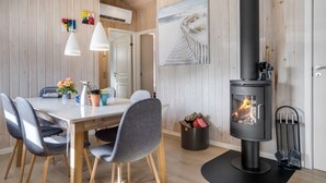 A modern kitchen with a central island, wooden panel walls, a black wood-burning stove, and a dining area with grey chairs and pendant lights.