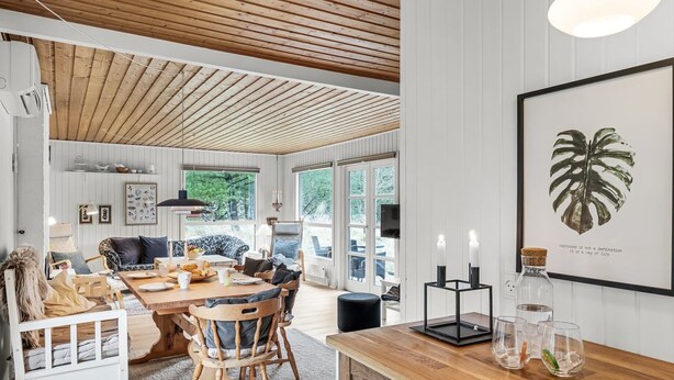 Un vaste espace de vie doté d’un plafond en bois, d’une table à manger avec des chaises, d’un canapé et d’un téléviseur. Un tableau encadré orne le mur, et une fenêtre offre une vue sur la verdure environnante.