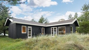 Une cabane moderne aux couleurs sombres, dotée d’un toit en pente, de grandes fenêtres et d’un espace verdoyant à l’avant.