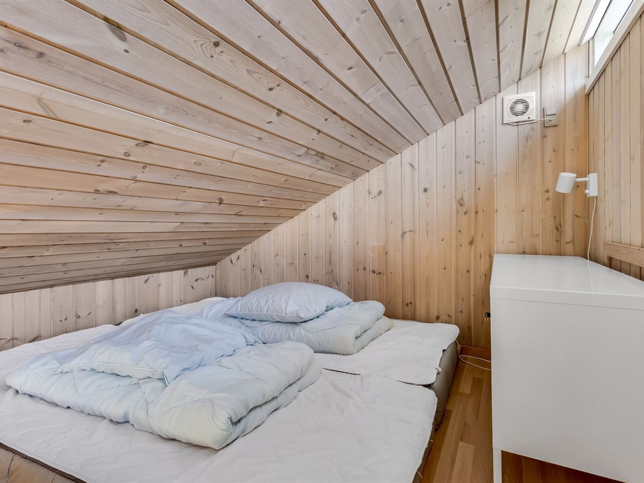 A small, wooden-paneled room with a sloped ceiling, a bed with white bedding, a white bedside table, and a wall-mounted light fixture.