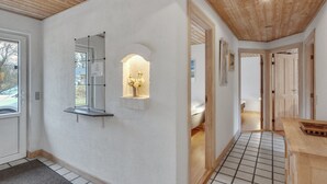 Un couloir avec un mur blanc, un plafond en bois, un sol carrelé, un miroir, une étagère avec un vase et une porte donnant sur une autre pièce.