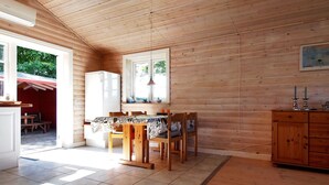 Una cabaña de madera con zona de cocina que incluye una mesa de comedor, sillas, un refrigerador y un ventanal con vistas al verdor.
