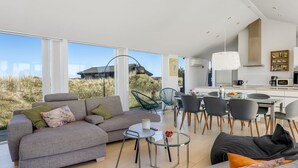 Una sala de estar moderna con un sofá gris, una mesa de centro de vidrio y un comedor con sillas, con una cocina al fondo.
