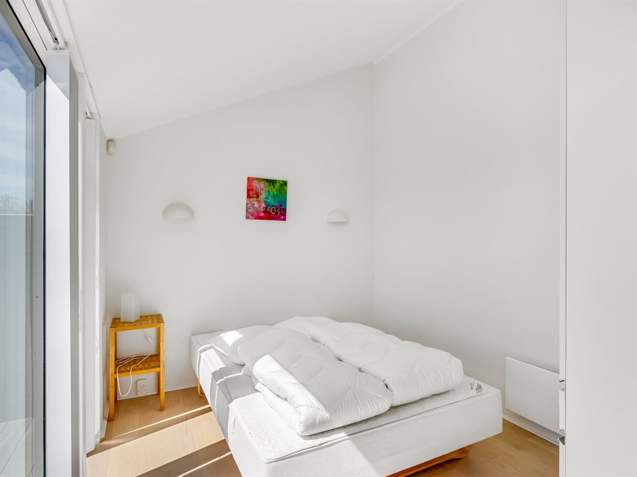 Een minimalistische slaapkamer met een bed, een klein nachtkastje, kunstwerk met felle kleuren en een raam.