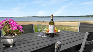 Pöydällä on viinipullo, kaksi lasia, lautanen täynnä hedelmiä ja ruukkuistutettu kasvi, ja taustalla avautuu näkymä järvelle.