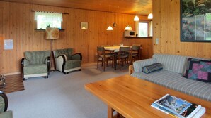 L’intérieur chaleureux d’un chalet avec des murs en bois, un coin repas, deux fauteuils, un canapé et une table basse en bois.