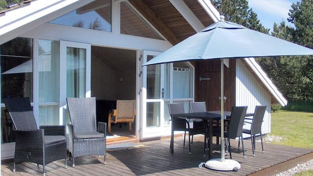 Een overdekt buitengebied met een tafel en stoelen, een grote parasol en uitzicht op een gebouw met grote ramen en een houten terras.
