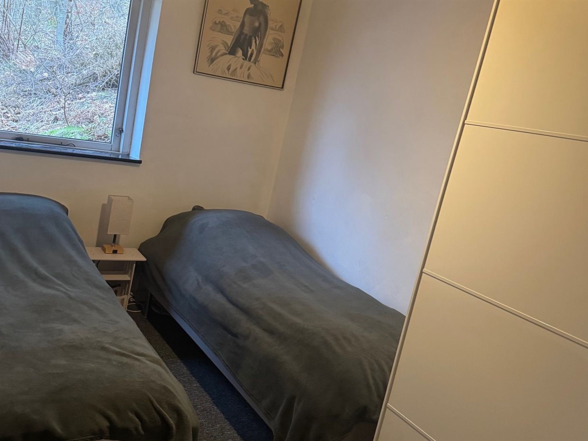 Un dormitorio con dos camas individuales, una mesita de noche, una lámpara y una ventana con vistas a árboles.