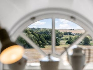 Une vue à travers une fenêtre à cadre blanc, qui révèle un paysage avec des arbres et une colline au loin.