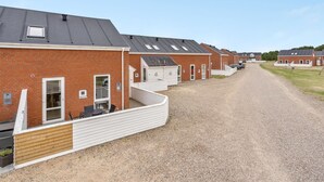 En række murstensbygninger med tilhørende udendørs siddeområder, en grusvej og en klar himmel.