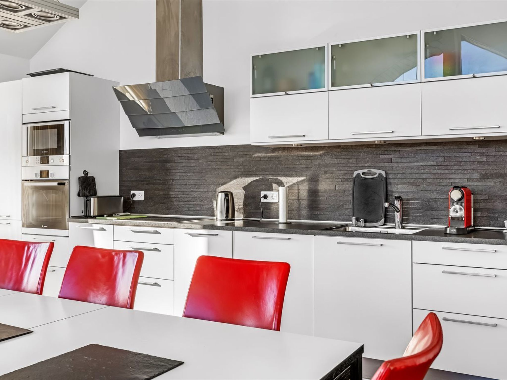 Une cuisine moderne avec des meubles blancs, un plan de travail noir et des chaises rouges.