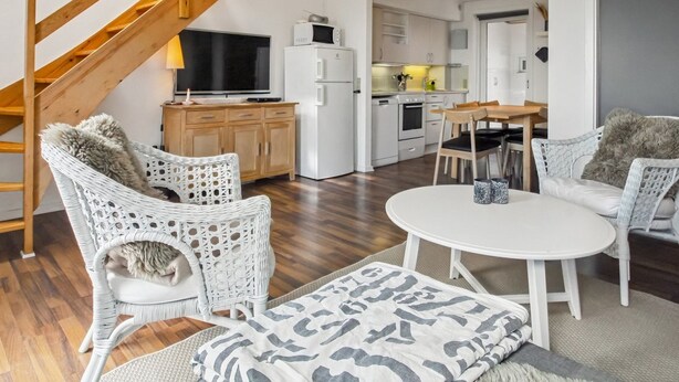 A modern living room with wicker furniture, a flat-screen TV, and a kitchen in the background.