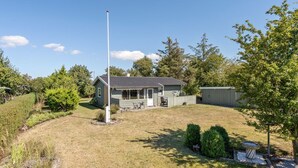 Ein eingeschossiges Haus mit Veranda, Fahnenmast und kleiner Gartenhütte in einem grasbewachsen Hof mit Bäumen.