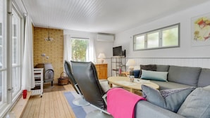 Ein gemütliches Wohnzimmer mit einer Couch, einem Sessel, einem Tisch und einem Fernseher. Eine Ziegelwand und ein Fenster mit Vorhängen sind ebenfalls zu sehen.