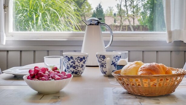 Ein Frühstücksbuffet mit einer Karaffe, Tassen, einer Obstschale und einem Korb mit Croissants auf einem Tisch neben einem Fenster mit Blick auf Grün.