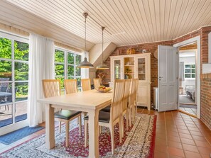 A dining area with a wooden table and chairs, a kitchen with a brick wall and cabinets, and a large window with curtains. - "Pauna" - 50m from the sea by Interhome (Væggerløse)