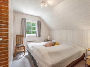 Ein Schlafzimmer mit schrägem Dach, einem fenster mit Vorhängen, einem Bett mit Kissen, einem Sessel und einem Nachttisch mit Lampe.