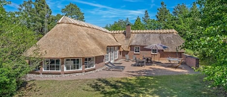 A thatched-roof house with a patio area, surrounded by trees.