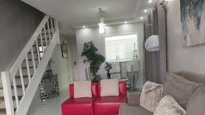 Uma sala de estar moderna com um sofá vermelho, um tapete felpudo cinzento e um sofá bege com almofadas fofas. Uma escada com grades brancas é visível ao fundo.