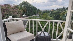 Uma varanda com cadeira e banquinho de vime, com vista para uma paisagem verdejante e uma cidade ao longe.