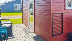 Ein rotes Holzgebäude mit Tür und Fenster, ein blaues Gebäude im Hintergrund und eine Terrasse mit Stühlen.
