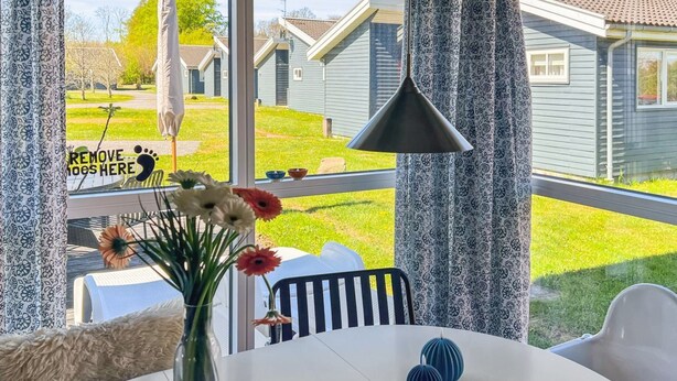 Ein Zimmer mit großem Fenster, weißem Tisch, Blumen in einer Vase und einer schwarzen Pendelleuchte.