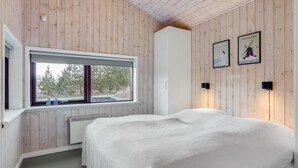 Slaapkamer met houten paneelwanden, een groot raam met uitzicht op bomen, een bed met een wit dekbed en twee wandlampen.