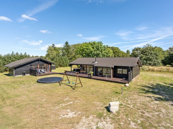 En moderne hytte med terrasse, trampolin og gyngestativ i en græsmark.