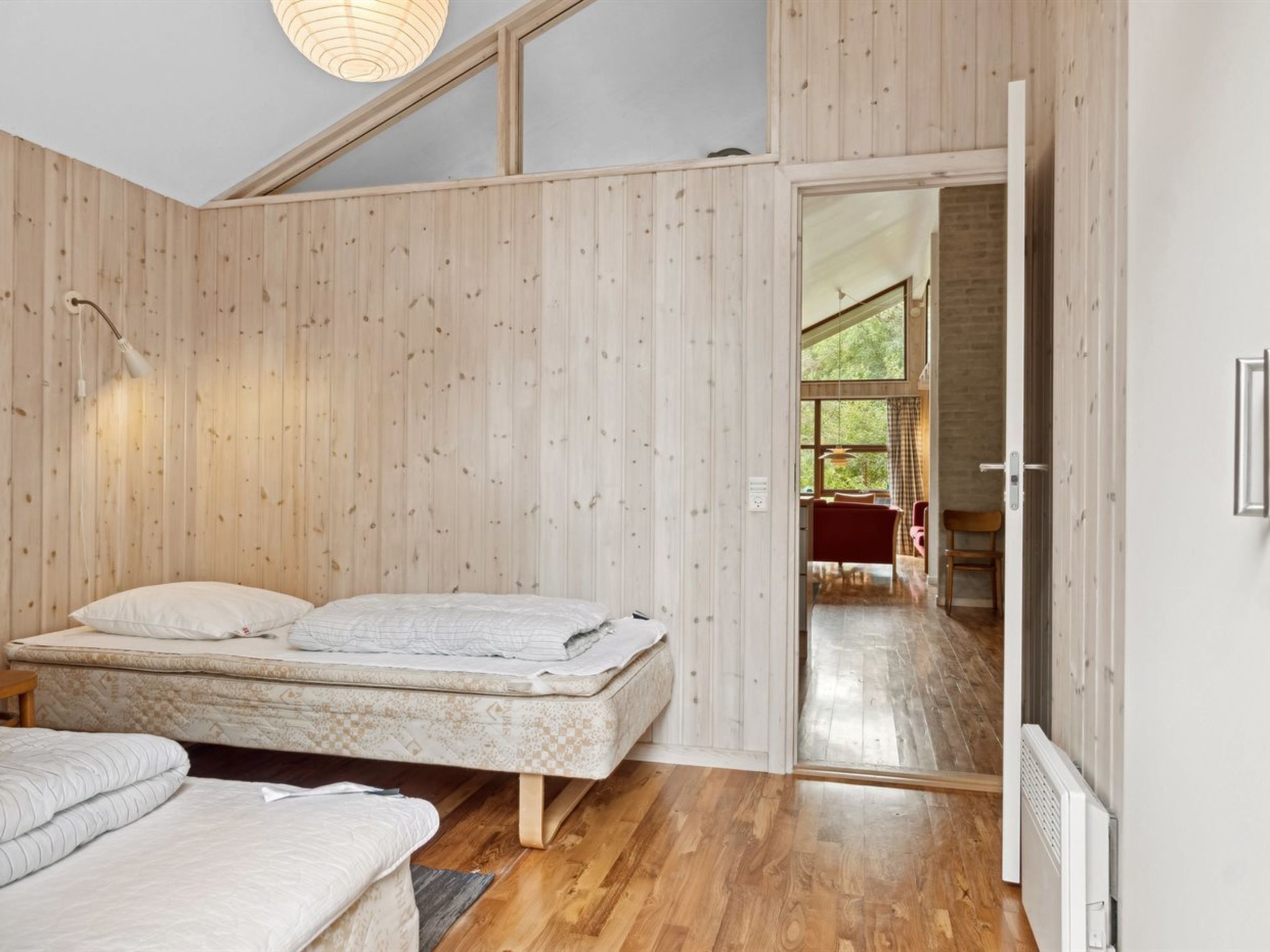 A bedroom with a wooden slatted wall, a bed, a small table, and a door leading to a bright room with a view of the outdoors.