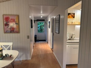 A hallway with a wooden floor, white walls, and framed pictures. There is a door at the end leading to a room with a window.
