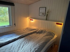 A single bed with a grey blanket in a room with white paneled walls and a window.
