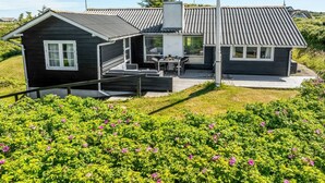 Een huis met een terras, een klein patio en een tuin vol roze bloemen.