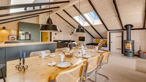 Un loft espacioso con techo alto, vigas de madera expuestas, una zona de comedor central con una mesa larga y un rincón de descanso acogedor con chimenea.
