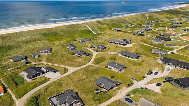 Vue aérienne d’une zone résidentielle côtière, avec des maisons, une plage et des éoliennes visibles au loin.