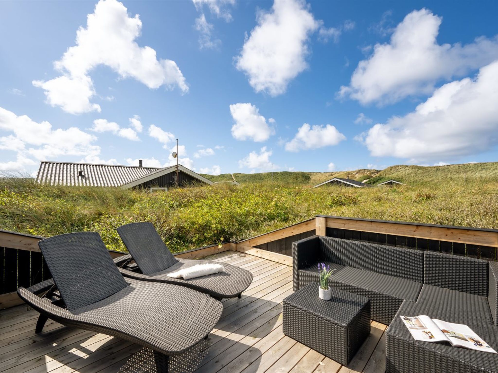 A deck with wicker furniture and a magazine, overlooking a grassy landscape with buildings in the distance.