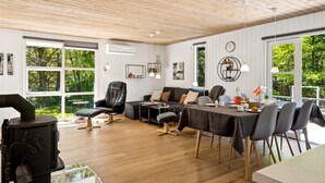 Amplia sala de estar con techo de madera, ventanas grandes con vista a zonas verdes, un sofá de cuero negro, una mesa de comedor con sillas y una estufa negra.
