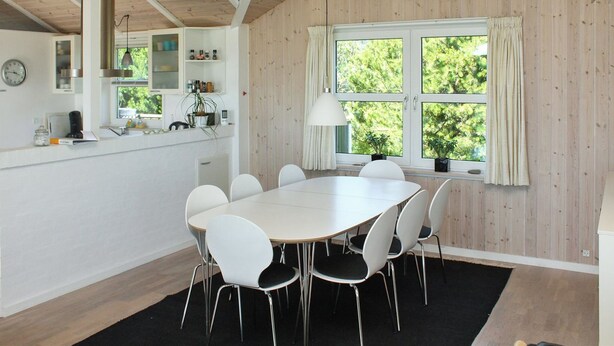 A modern kitchen with white cabinets, a round dining table, and six chairs. There is a window with curtains, a clock on the wall, and a plant on the windowsill. - "Calina" - 300m from the sea by Interhome (Sæby)