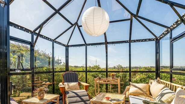 A glass-enclosed room with a wooden deck, furnished with a sofa, chairs, and tables, overlooking a vineyard.
