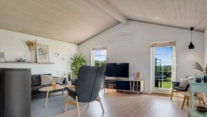 A spacious living room with a wooden ceiling, a grey armchair, a TV, and a dining area with a table and chairs.